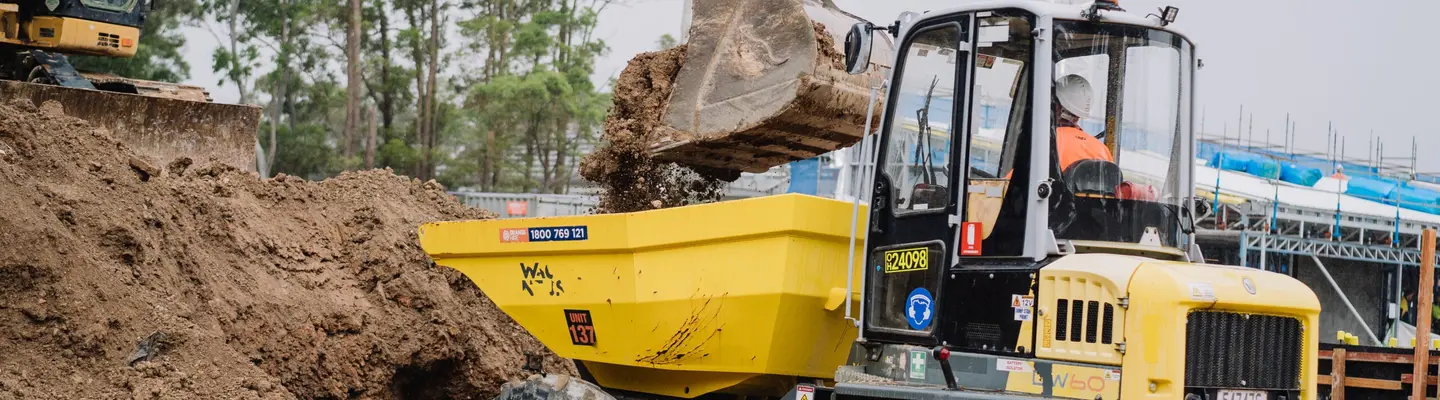 Man operating an excavator with attachment digging soil at a construction site with safety barriers in the background.