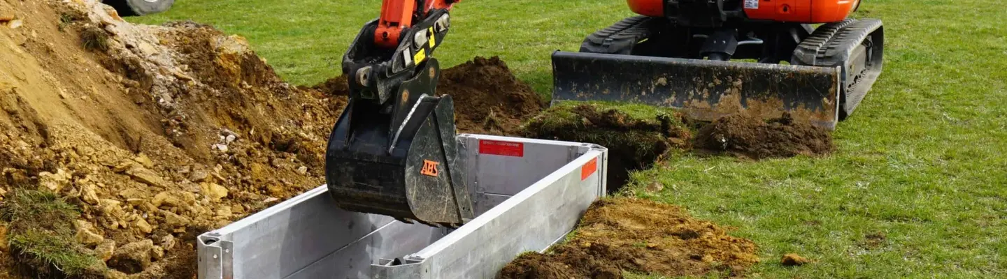 Worker operating excavation machinery near braced trench with visible hazardous material signs on a construction site.