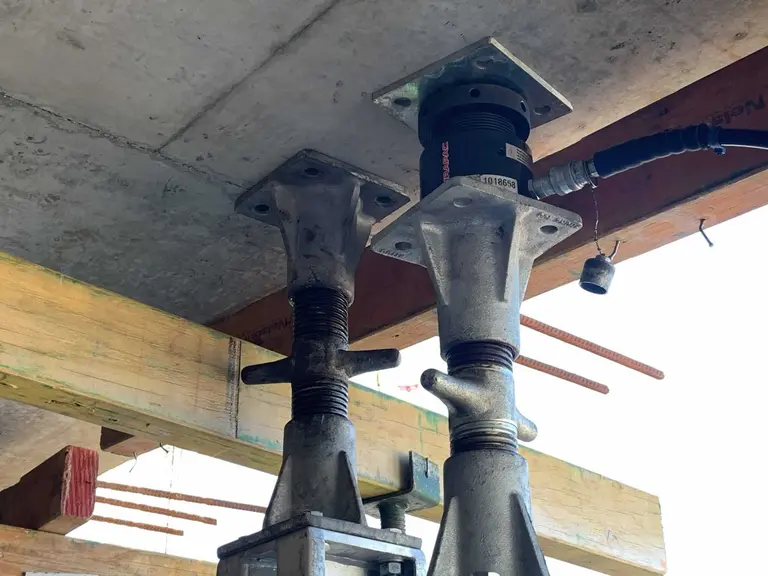 A close-up of adjustable steel props supporting a concrete slab, with a hydraulic jack installed on one support beneath a timber formwork structure.