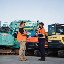 Two workers in uniform smiling while standing in front of construction equipment at a job site.