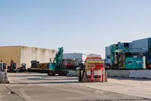 Construction site with various equipment, a water truck spraying dust control, and water-filled safety barriers in place.
