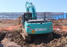 Excavator digging at an active construction site, with safety barriers and warning signs visible around the work area.