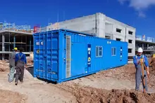 Blue shipping container office installed on a construction site with workers nearby.