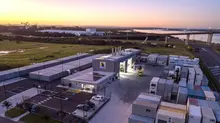 Aerial view of a container facility and warehouse complex beside a river and bridge at sunset.